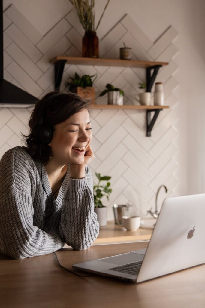 Young cheerful female smiling and talking via laptop while sitting at wooden table in cozy kitchen