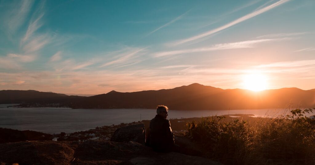 A person sits on a hilltop, watching a vibrant sunset over a mountain and lake landscape.